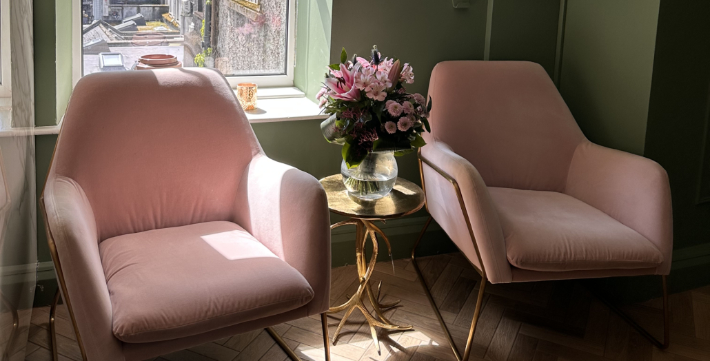 two pink armchairs with gold table in between them. Pink flowers on table. Sunny window in background