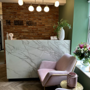 Odeon Room reception area. Brick wall with light fixture on roof. Green window with pink chairs in front of it. Flowers and plants on desk and window sill.