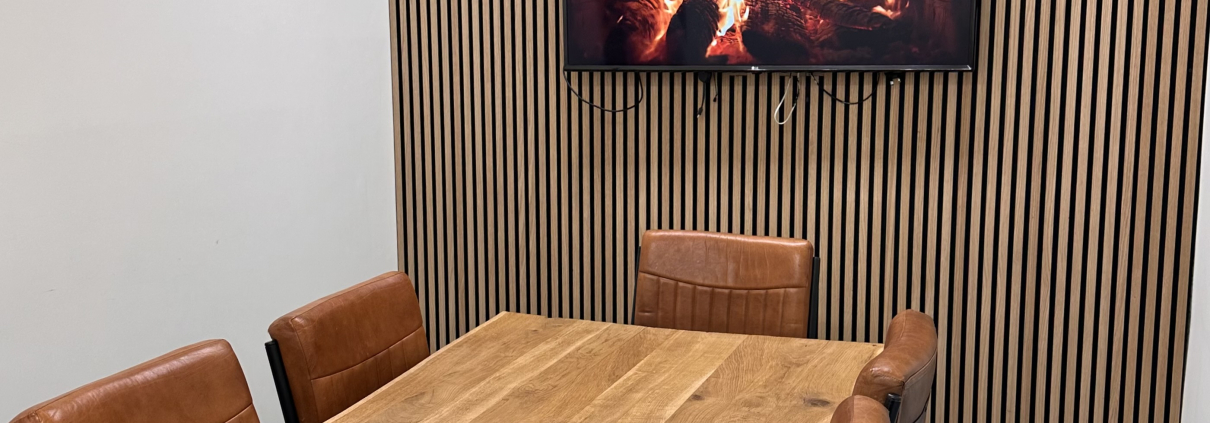 Office boardroom with wood pannelling feature on back wall, a wooden oak table with brown chairs and a picture of a fire on a tv mounted on the wall.